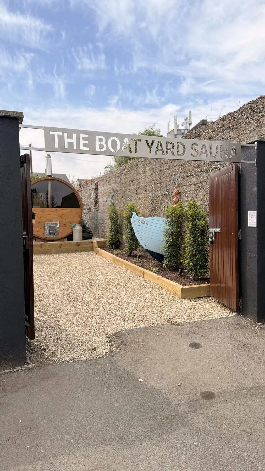 The Boat Yard Sauna entrance gate, a barrel sauna and blue boat planter beyond