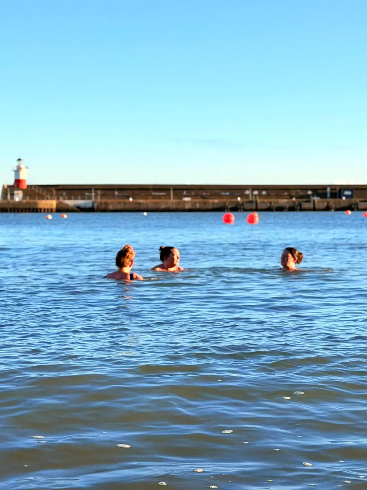 Three friends sea-swimming together by the harbour wall and lighthouse